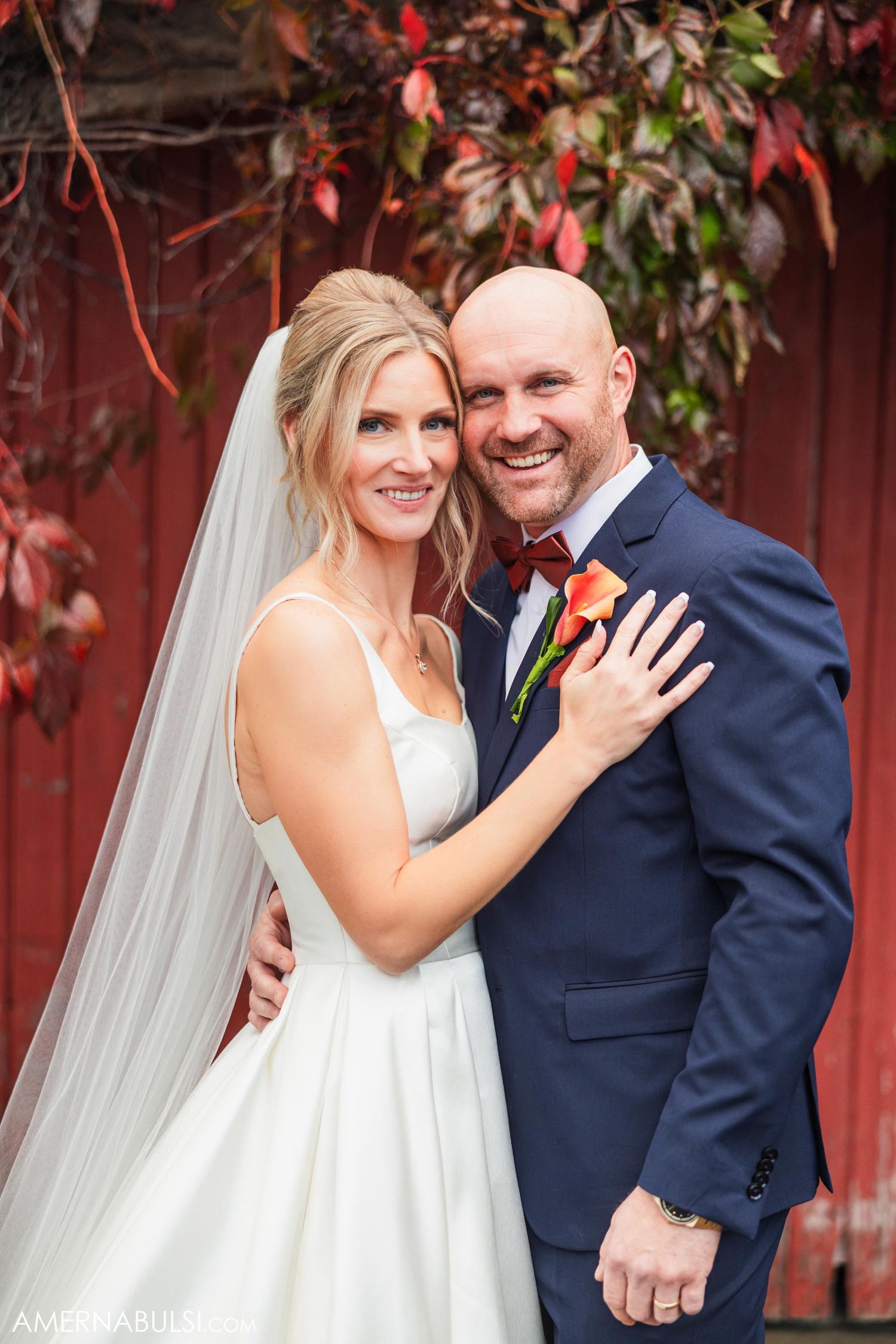 Wedding couple posing against red barn with autumn leaves and rustic backdrop.