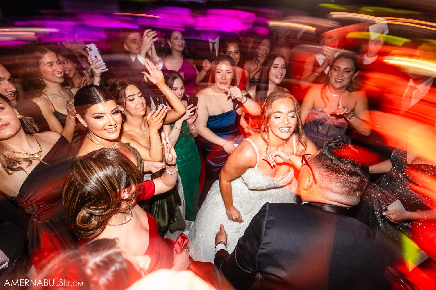 Wedding party guests dance energetically under vibrant purple and orange lighting on a crowded dance floor.