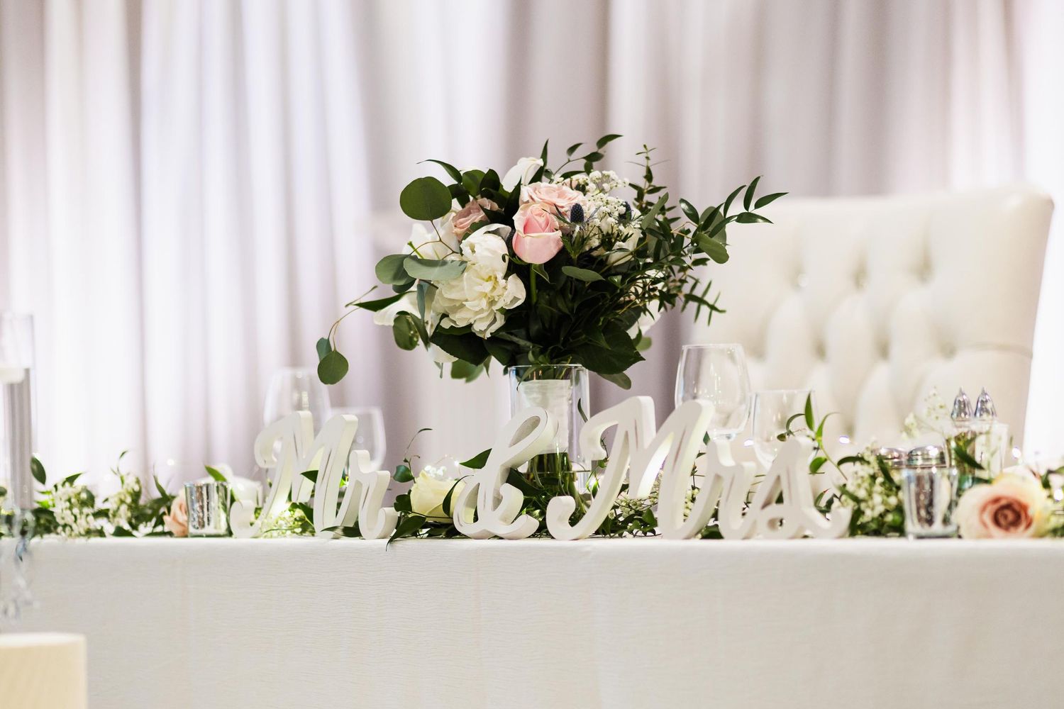 Wedding head table decorated with white roses and greenery against an elegant tufted backdrop.