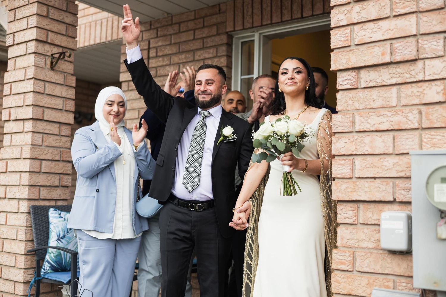 Newlyweds celebrate with raised arms while guests cheer outside a brick building.