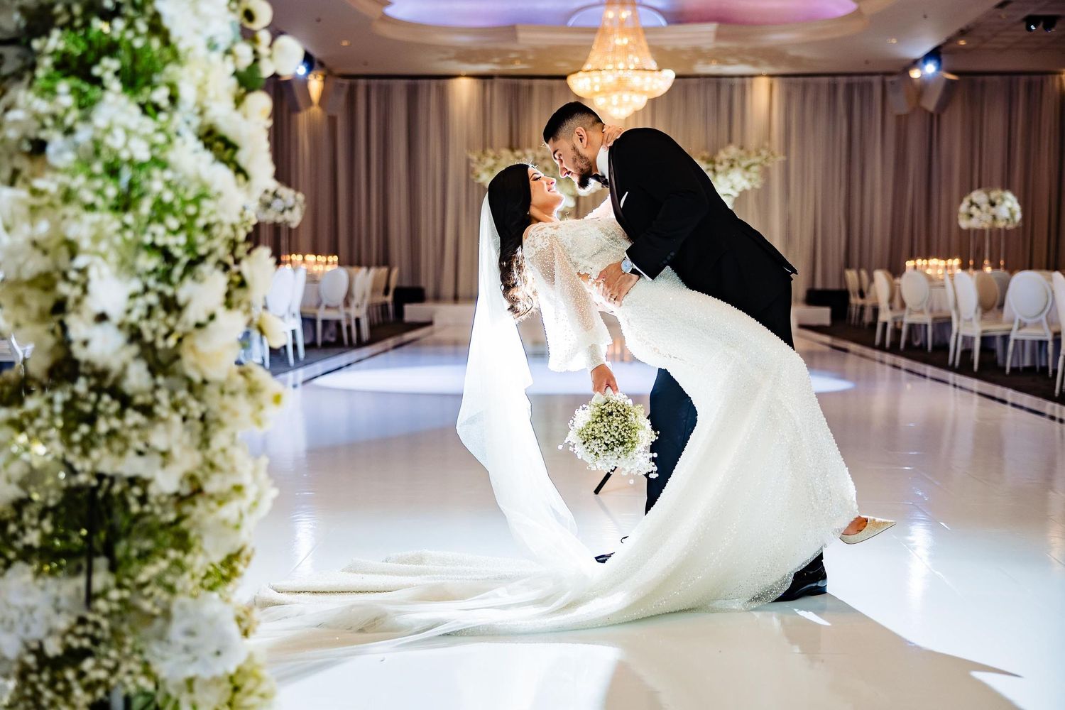 A romantic wedding dip pose on an elegant white dance floor surrounded by floral arrangements and chandeliers.