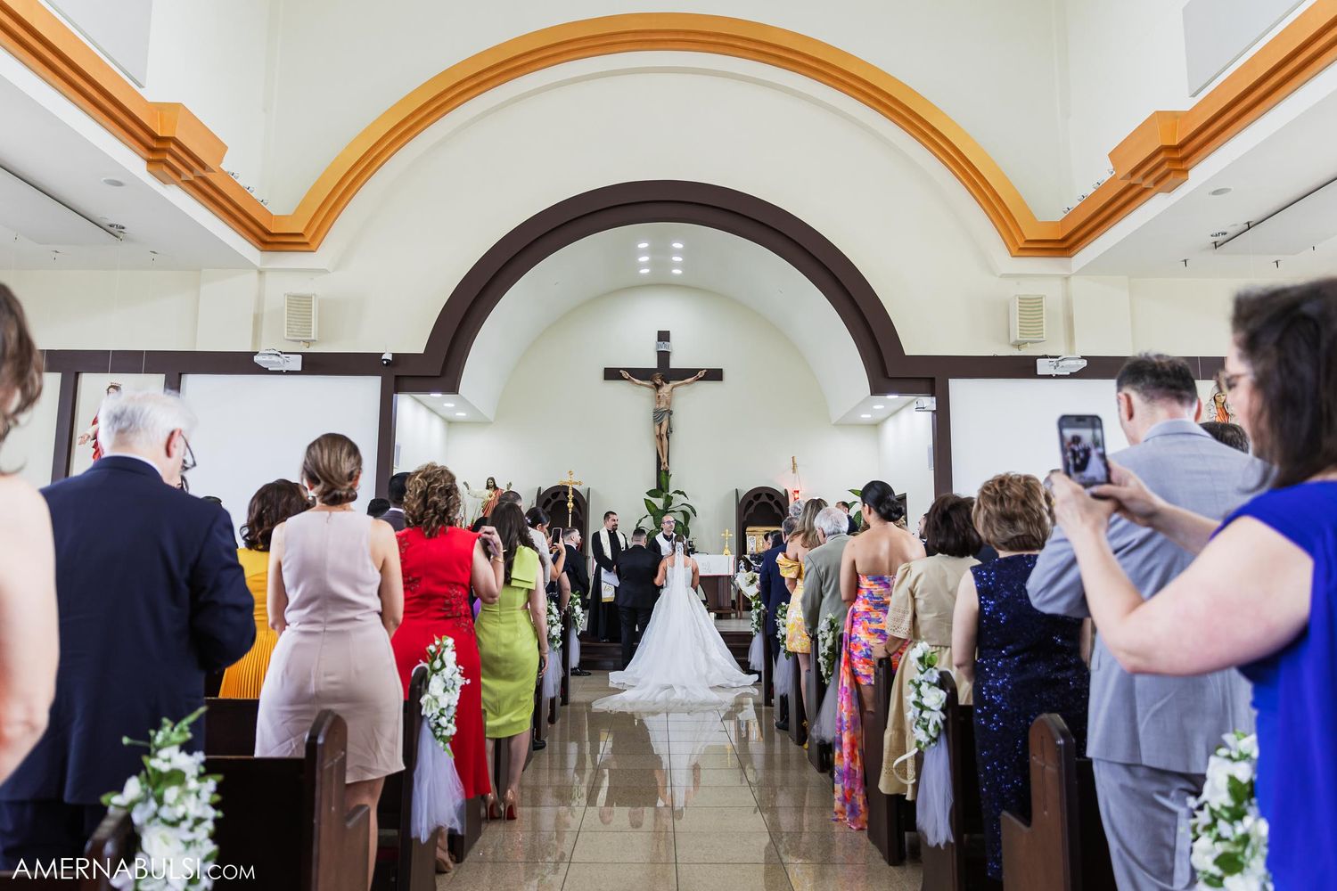 Wedding ceremony in progress down a long church aisle with guests seated on both sides.