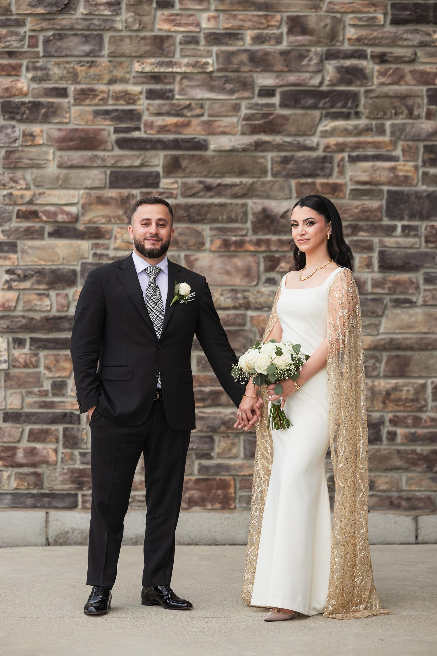 Formal wedding portrait against textured stone wall with white floral bouquet.
