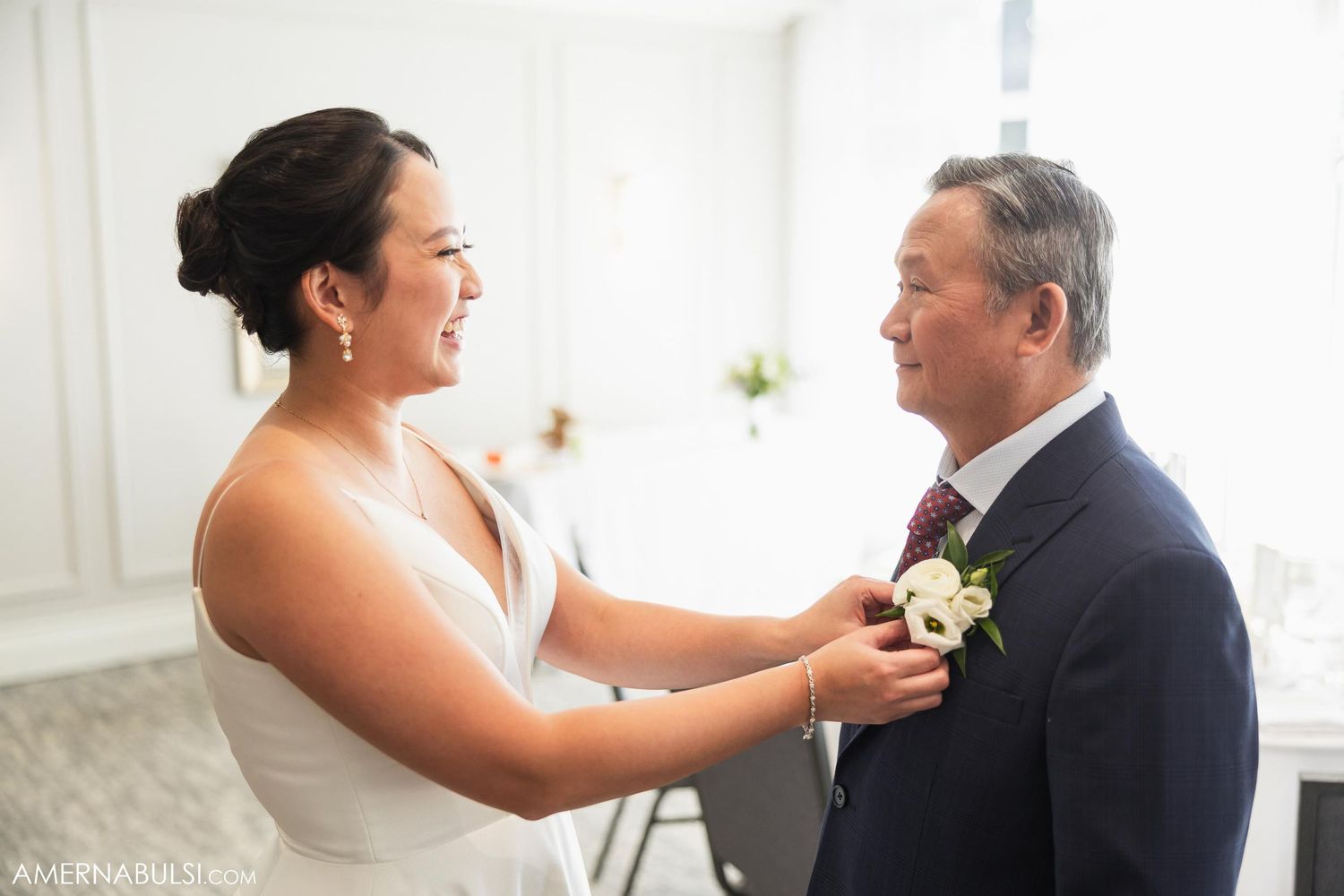 A touching moment as a bride helps adjust a boutonniere on her father's suit jacket in a bright indoor setting.