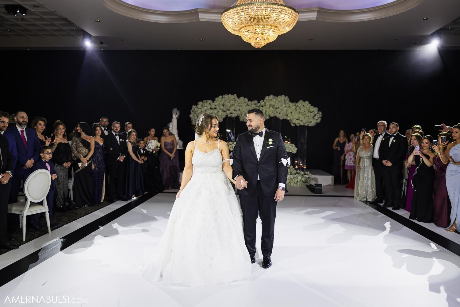 Newlyweds walk down a white aisle under a crystal chandelier as guests in formal attire look on.