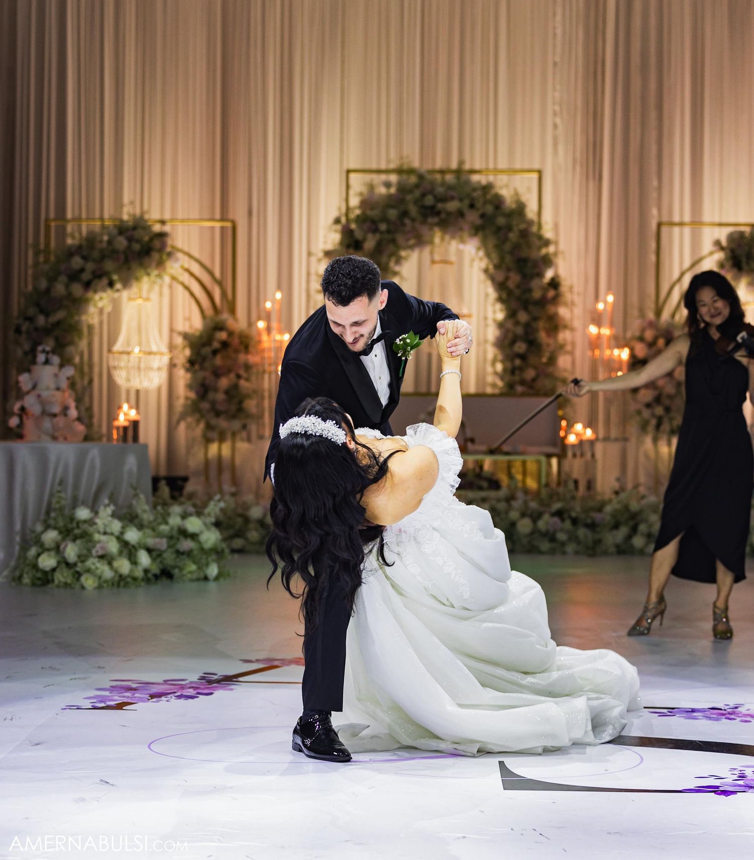 A couple performs a dramatic dip during their first dance at an elegant wedding reception with floral arches.