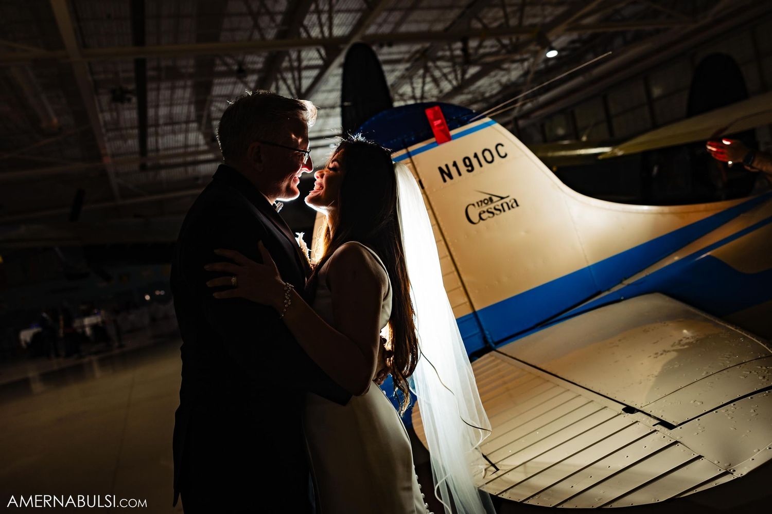 Couple sharing intimate moment next to vintage aircraft in dimly lit hangar.