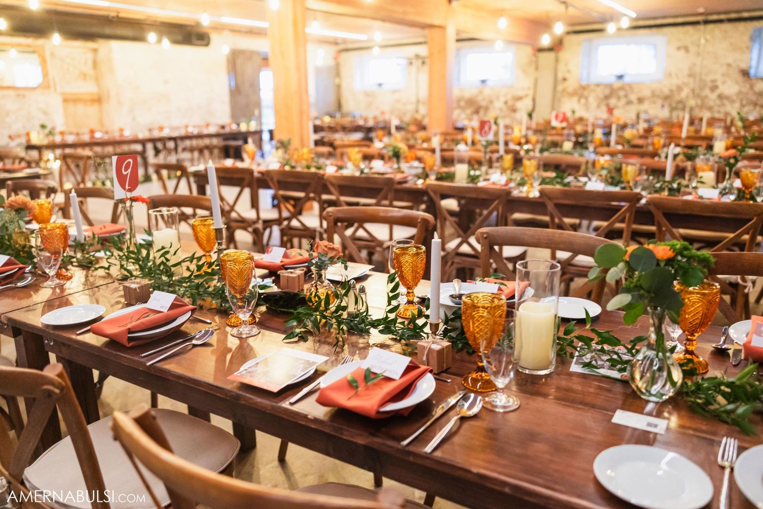 Rustic barn wedding reception tables set with greenery, glassware and wooden cross-back chairs.