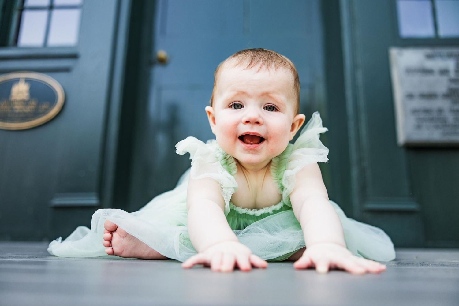 Smiling baby in mint green dress lies on gray floor in front of dark blue door.