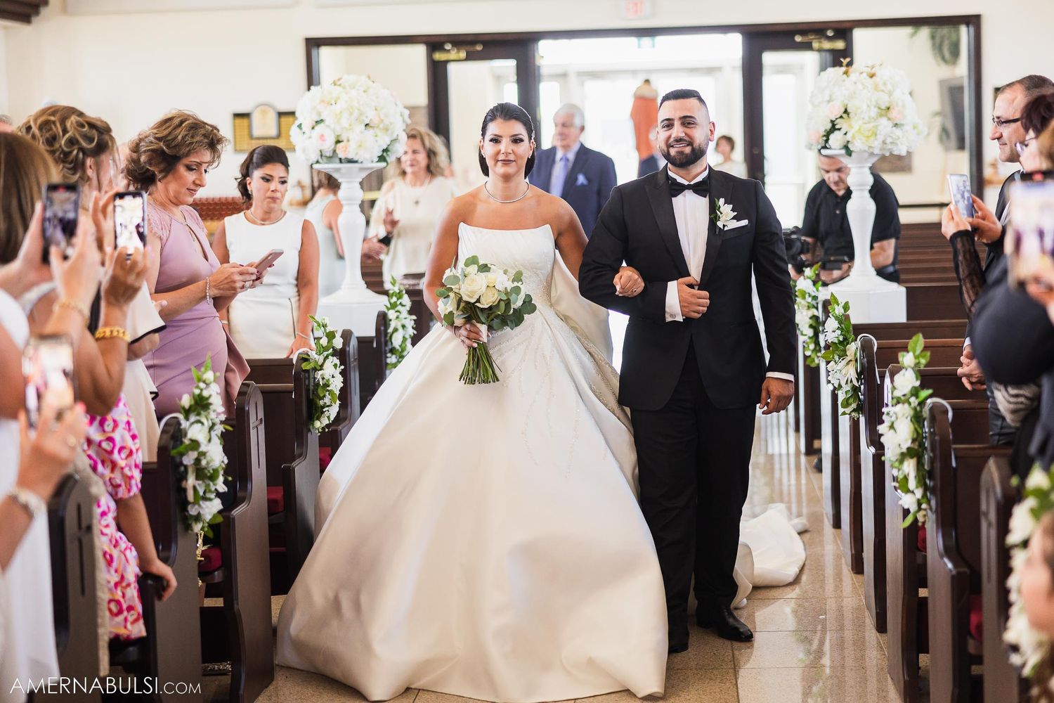 A wedding couple processes down a church aisle decorated with floral arrangements.