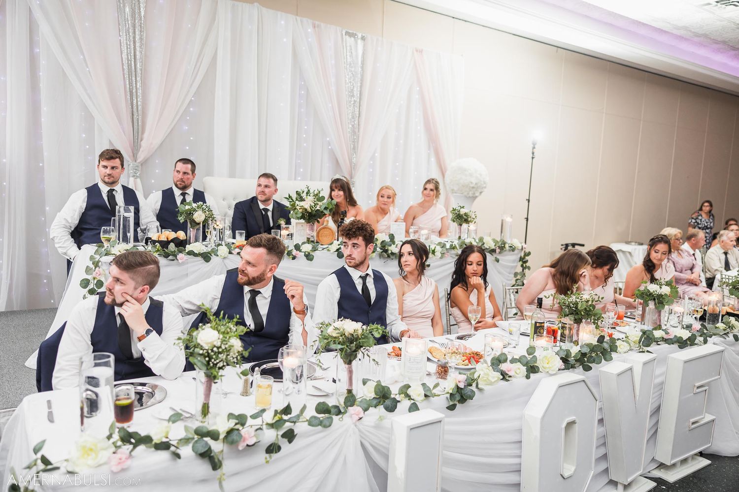 Wedding party seated at an elegantly decorated head table with white floral arrangements.