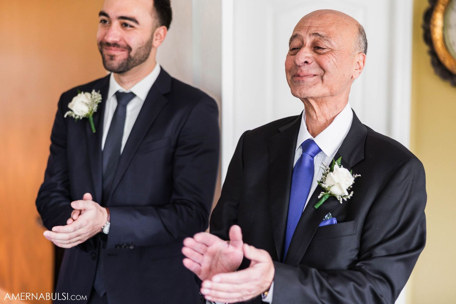 Two men in dark suits with boutonnières share a lighthearted moment during wedding preparations.