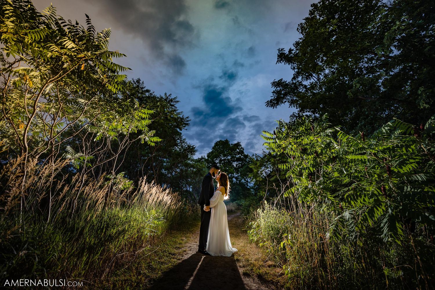 Couple standing on pathway surrounded by lush greenery under dramatic stormy sky.