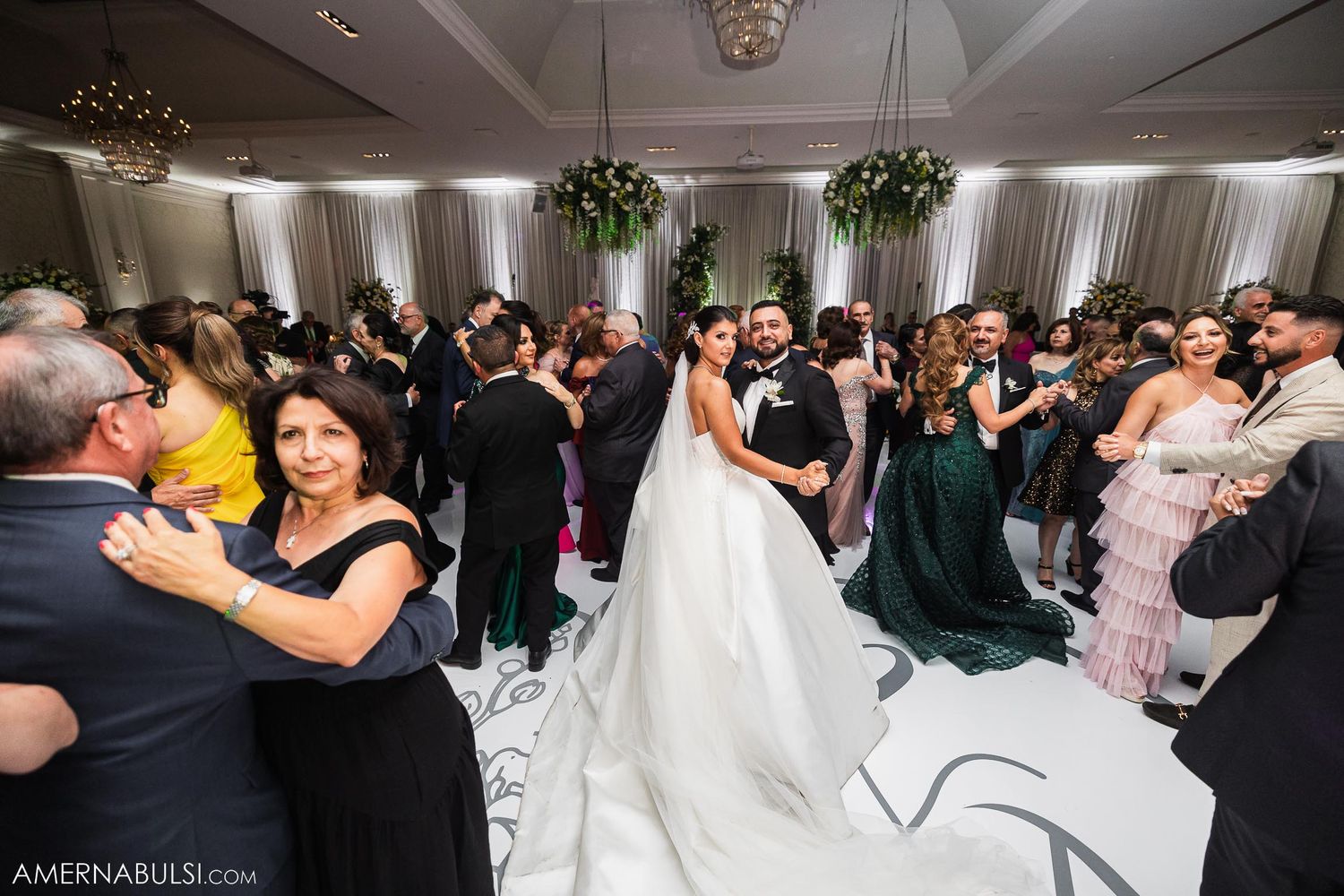Wedding guests dancing and celebrating on an elegant reception dance floor with white and floral decor.