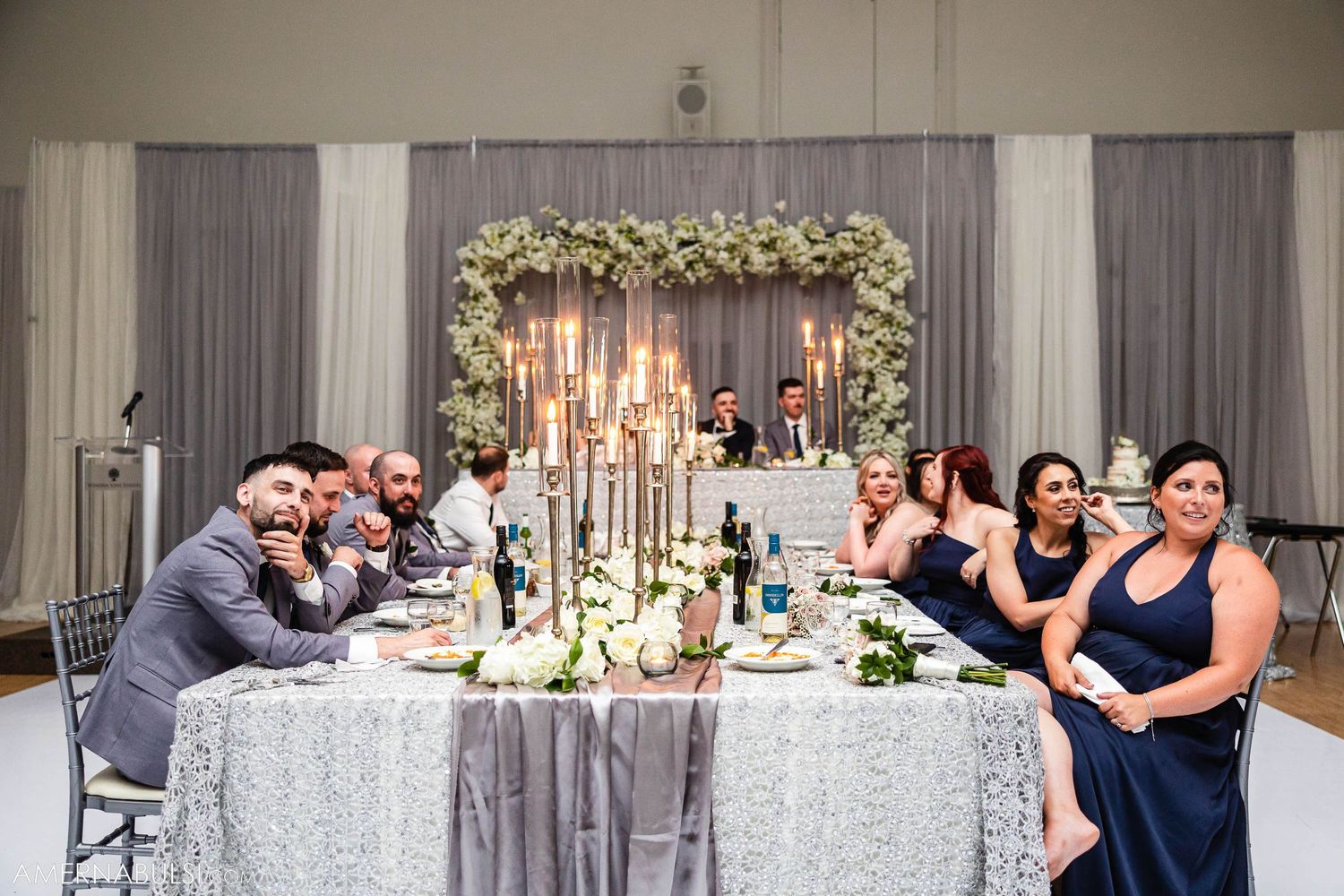 Wedding party seated at an elegantly decorated reception table with floral arch backdrop.