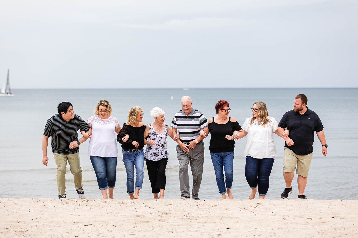 A group of people walk together arm in arm along a sandy beach with a sailboat visible in the distance.