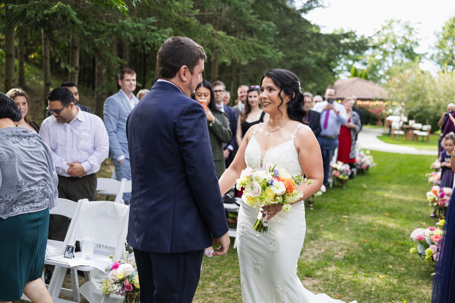Bride smiling at groom during outdoor wedding ceremony with guests seated on white chairs among lush green trees.