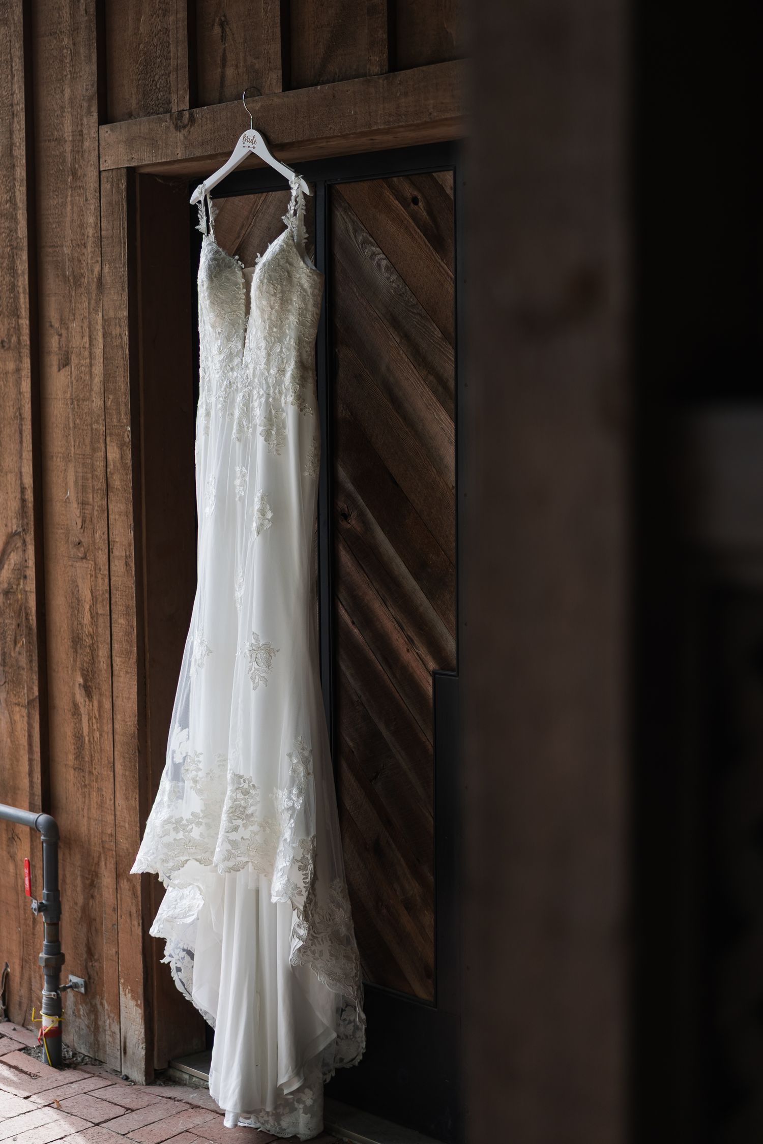 Elegant lace wedding dress hanging on a wooden barn door, showcasing intricate floral details and flowing skirt.
