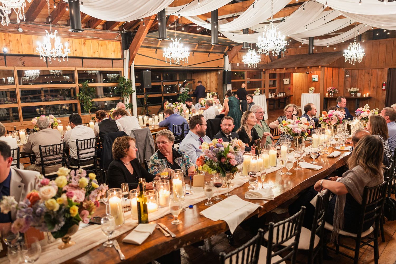 Guests gathered at long wooden tables during an elegant rustic barn wedding reception with chandeliers and floral centerpieces.