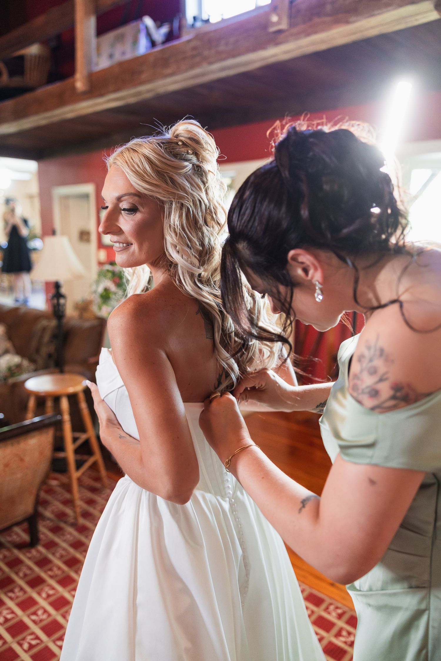 Bridesmaid with tattoos fastening buttons on back of bride's white wedding dress in rustic barn.