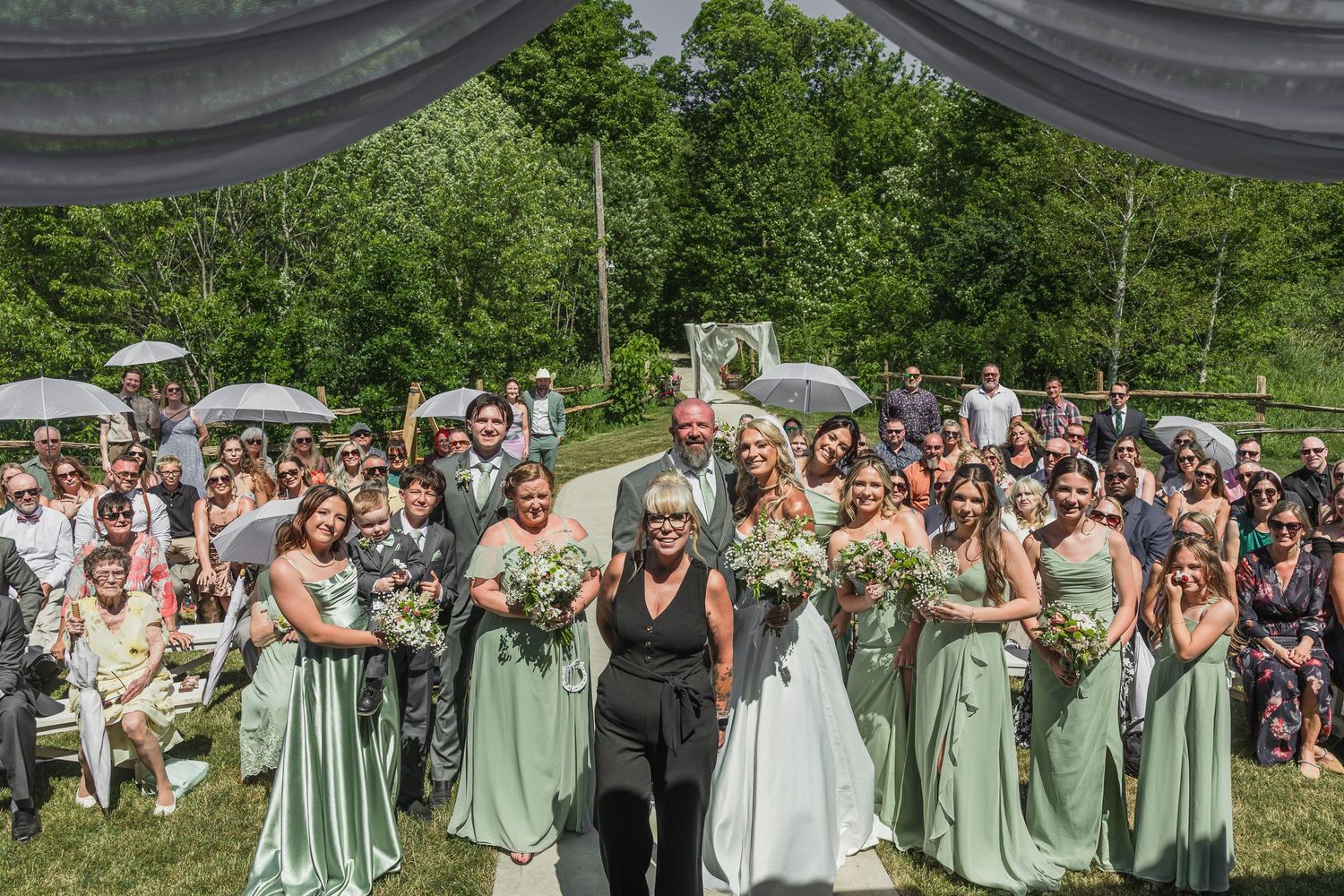 Bride and bridesmaids in sage green dresses walking down outdoor aisle with wedding guests gathered around.