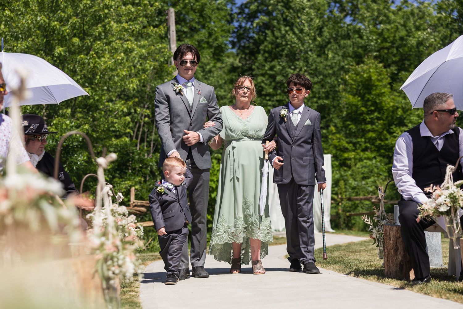 Wedding procession with family walking down outdoor aisle, woman in green dress flanked by two men in gray suits.