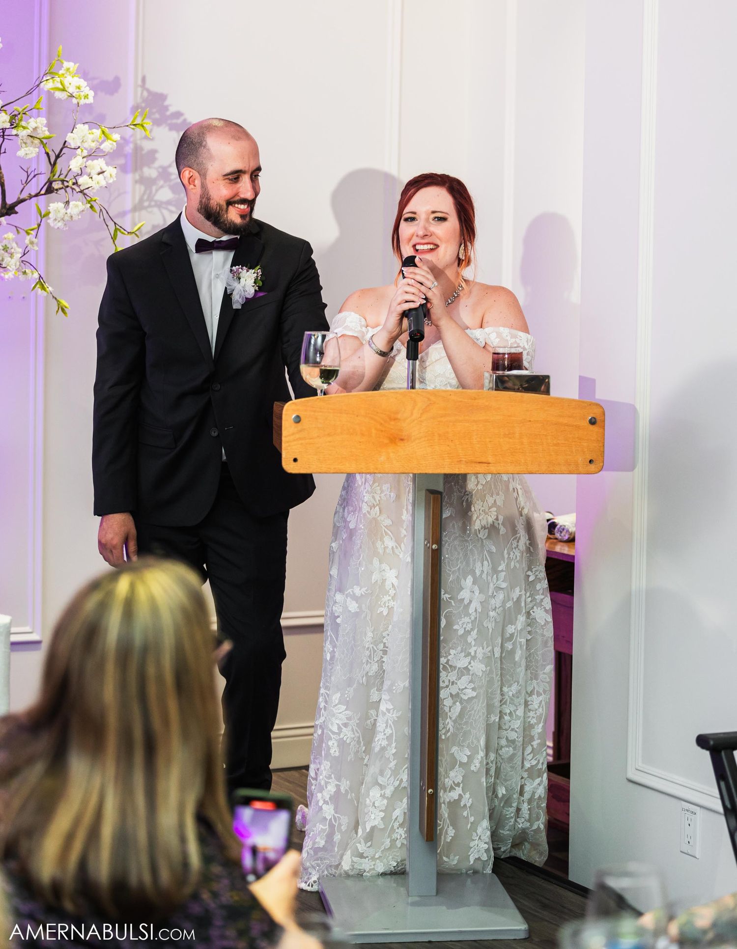 Couple giving speech at wooden podium during wedding reception with purple floral decor.
