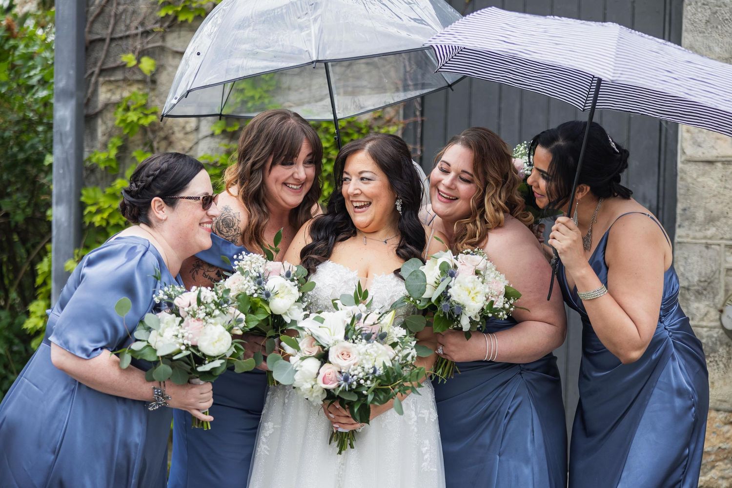 Wedding party shares laughs while holding umbrellas and white bouquets.