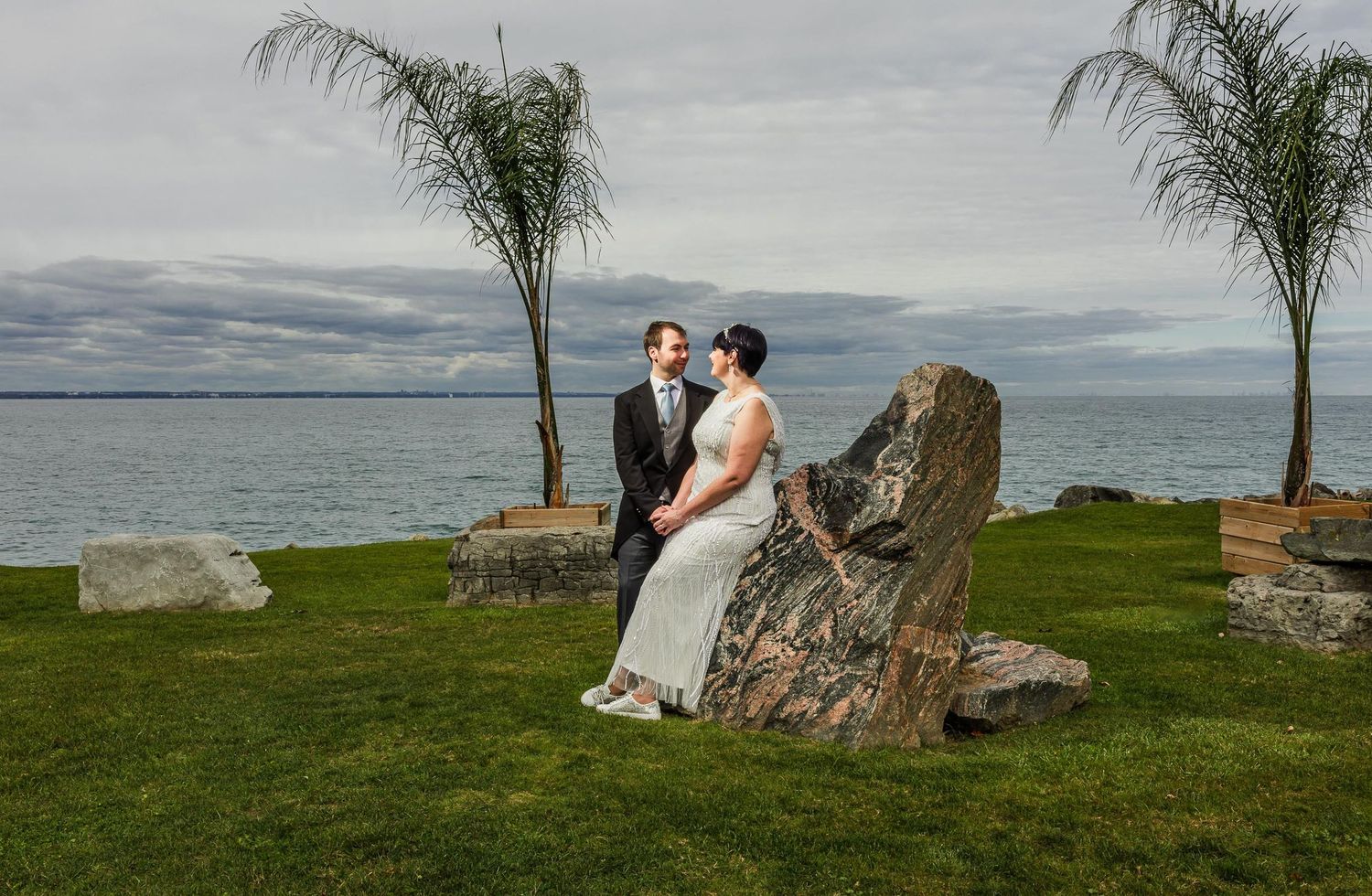 Wedding couple poses by large rocks and palm trees overlooking a serene ocean view.
