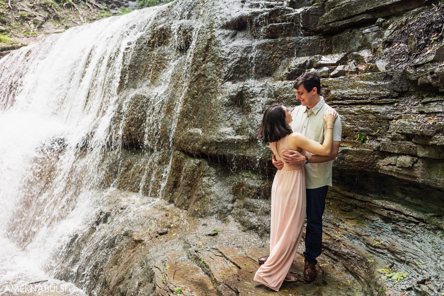 Romantic embrace near a flowing waterfall with rocky cliffs in the background.