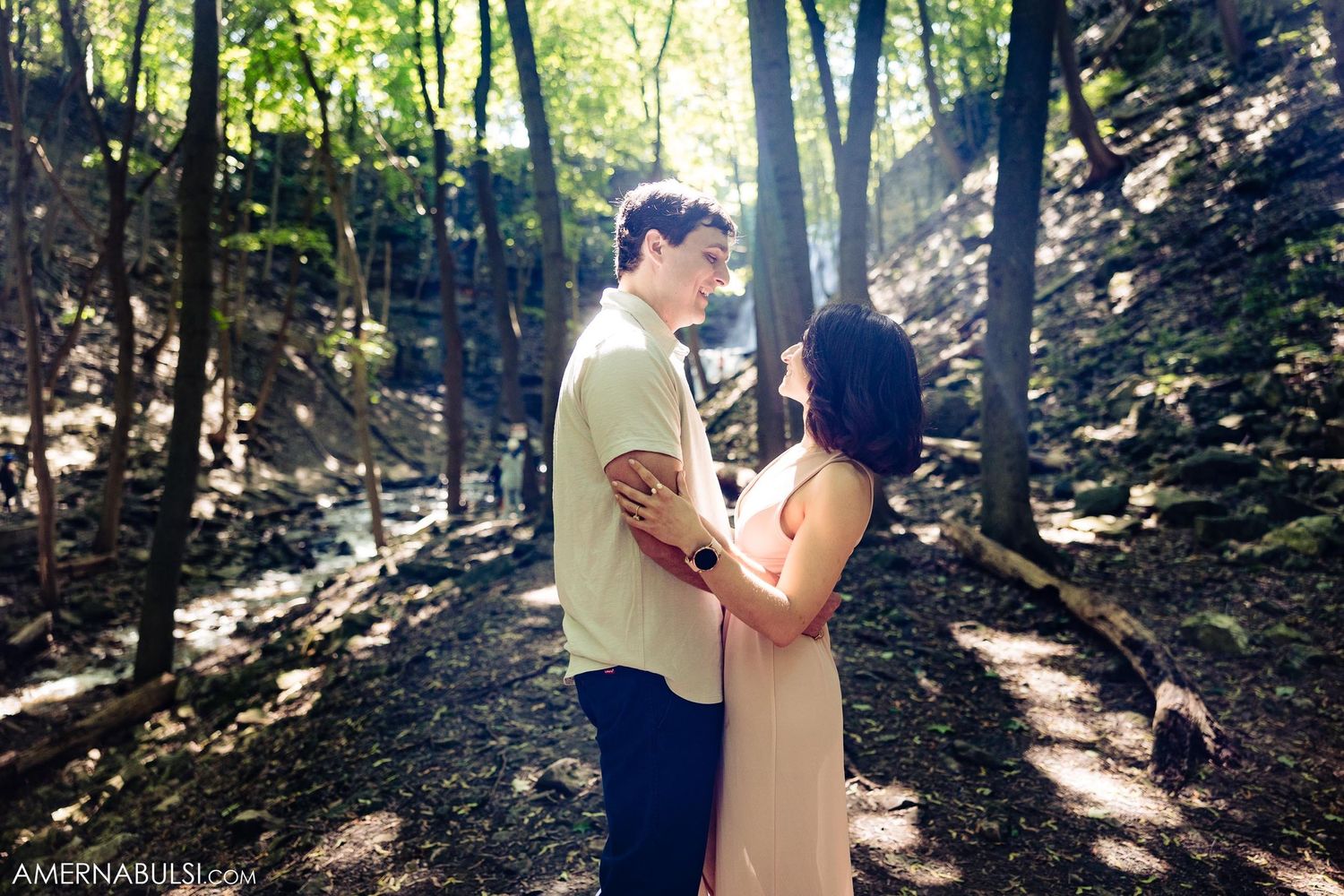 A couple shares an intimate moment on a sunlit forest path surrounded by trees.