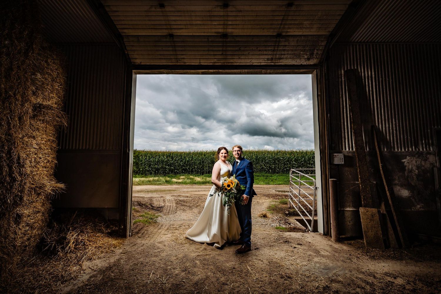 Wedding scene framed within rustic barn doorway overlooking cornfield under moody sky.