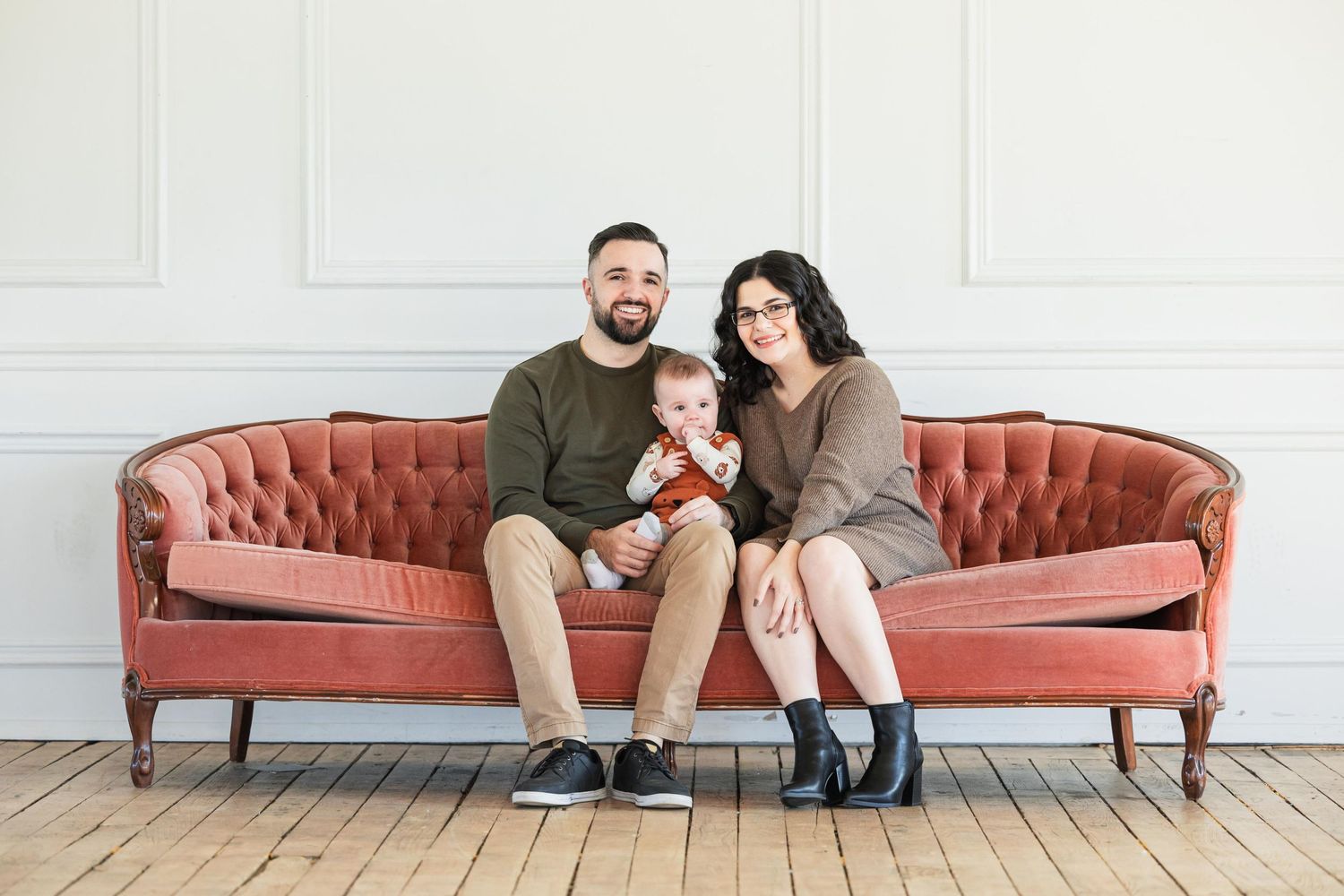 A family sits together on a vintage pink velvet sofa against a white wall.