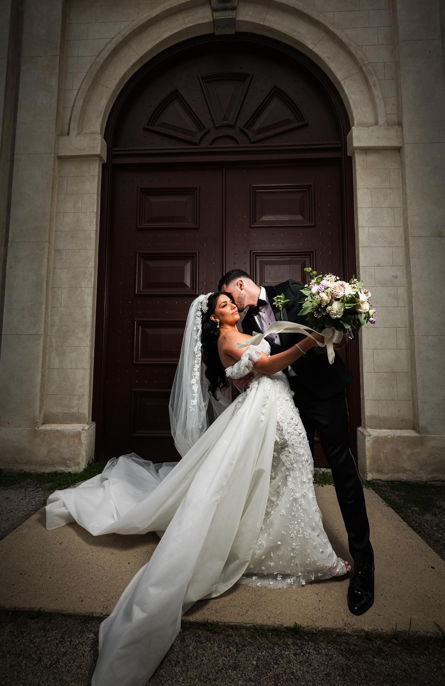 Elegant wedding couple poses dramatically in front of grand arched wooden door.