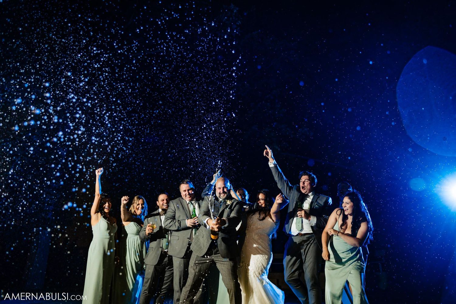 Wedding group poses triumphantly as confetti rains down under blue spotlights.