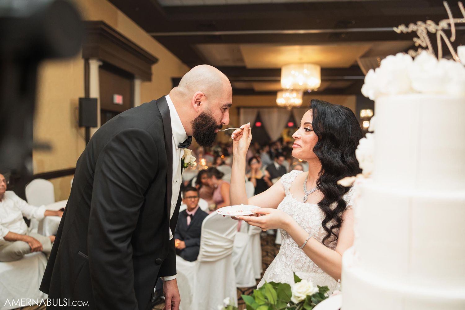 Bride and groom playfully feeding each other wedding cake while guests watch and smile.