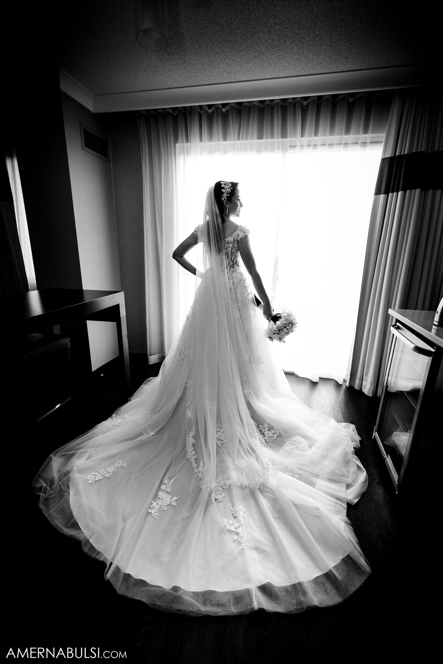 Dramatic black and white silhouette of bride in lace wedding gown with cathedral train by window.