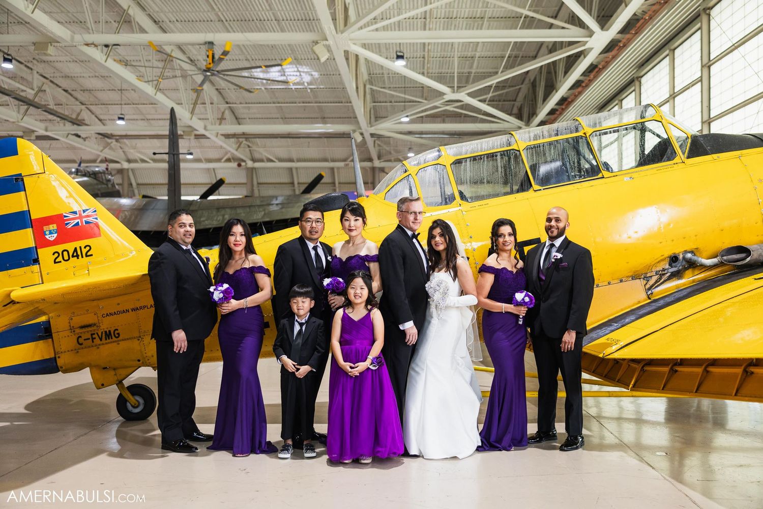 A wedding party in purple and black attire poses in formation beside a yellow vintage aircraft.