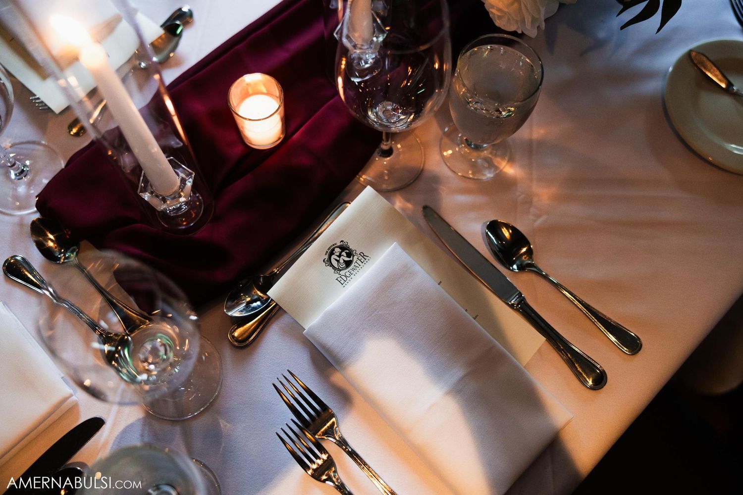 Formal table setting with white linens, burgundy accents, and glowing candles.