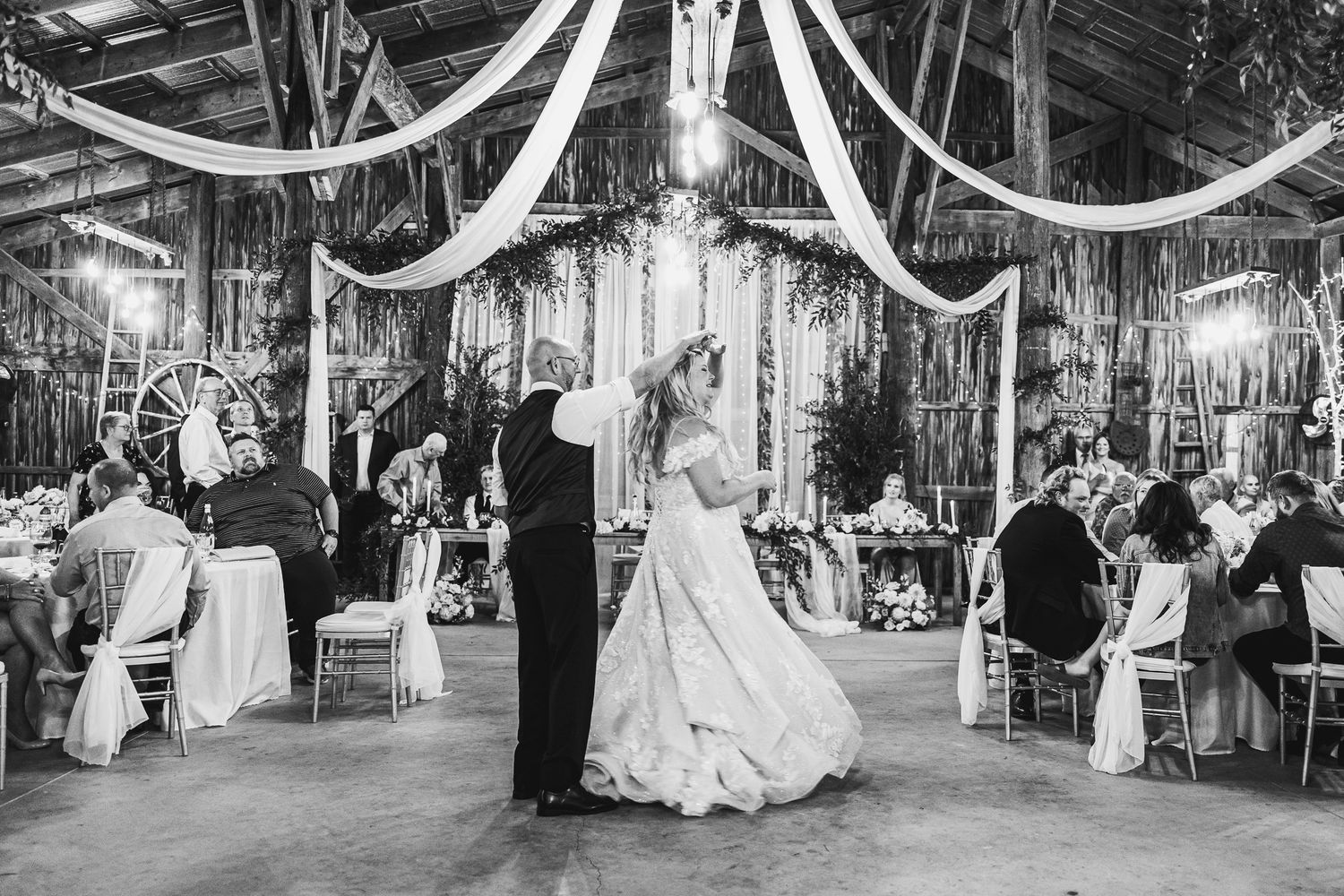 Elegant wedding reception with white fabric draping and crystal chandeliers in a rustic barn venue.