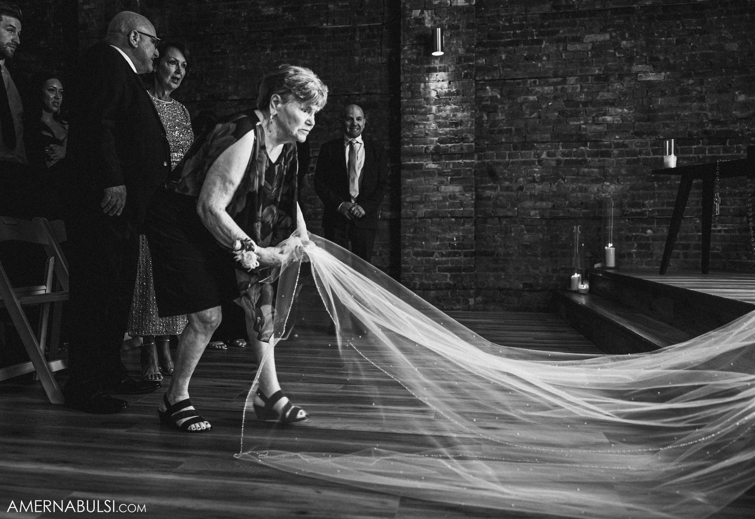 Black and white image of wedding veil being arranged on dance floor in brick-walled venue.