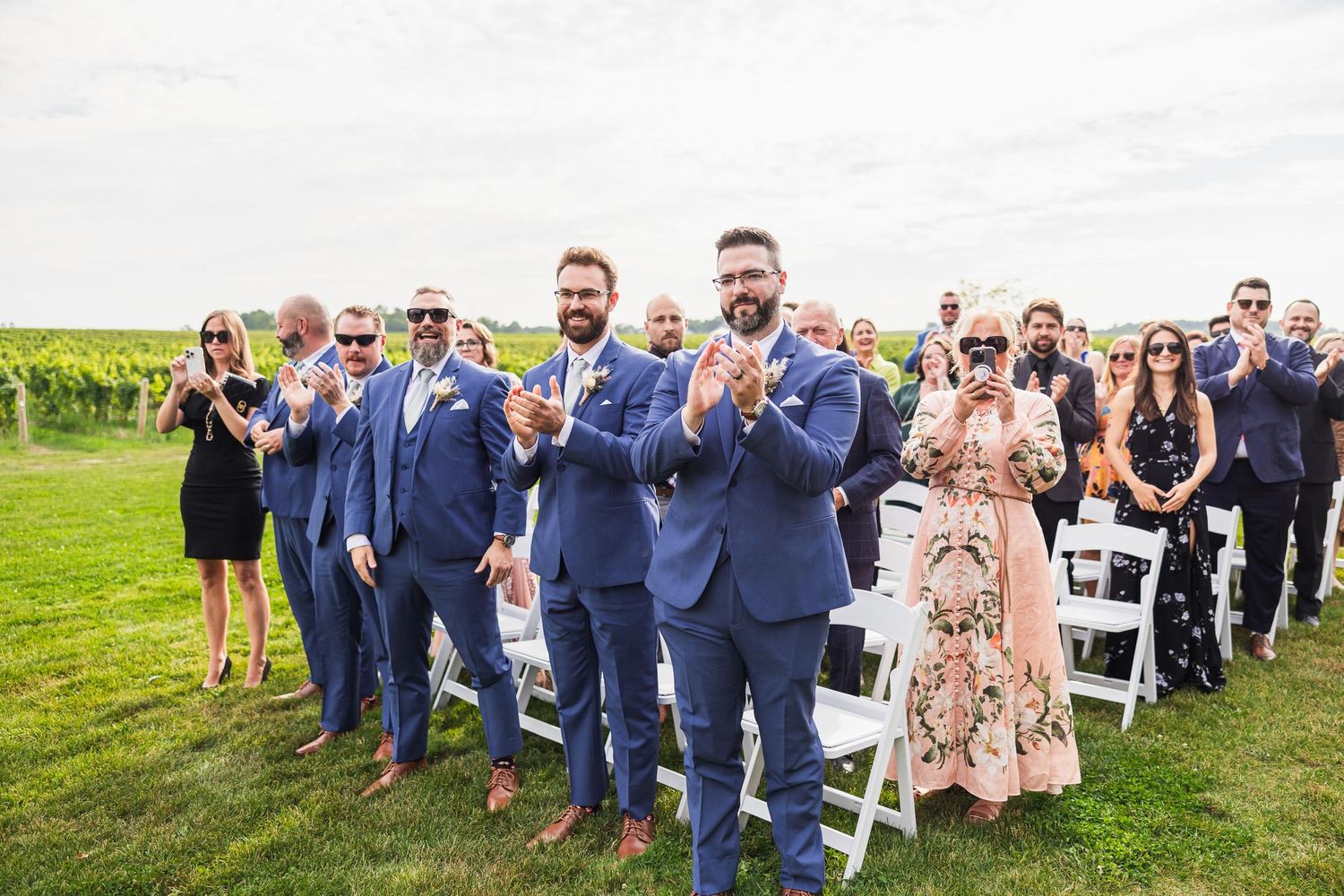 Wedding guests in blue suits and formal attire applauding during outdoor ceremony.