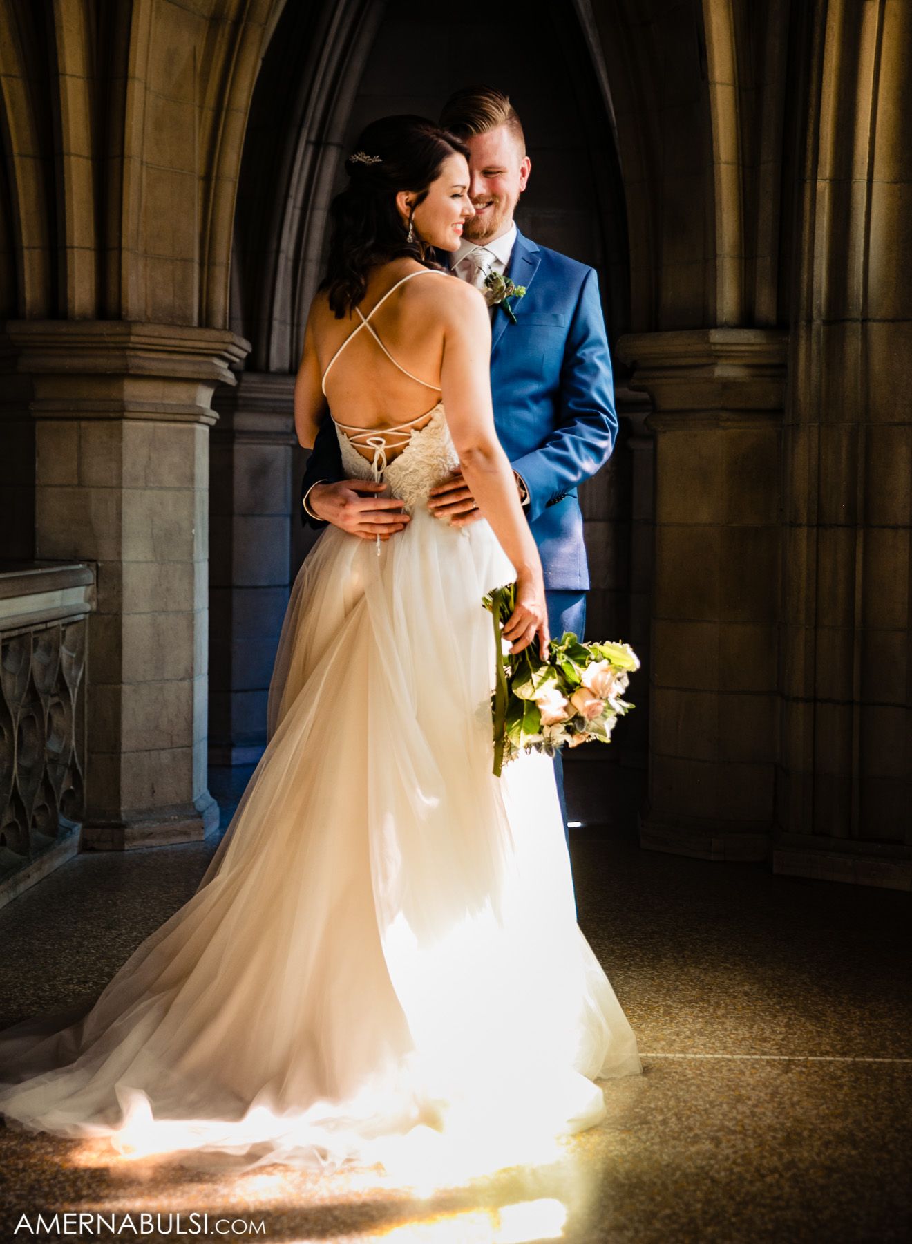Bride in flowing white dress illuminated by natural light in stone archway.