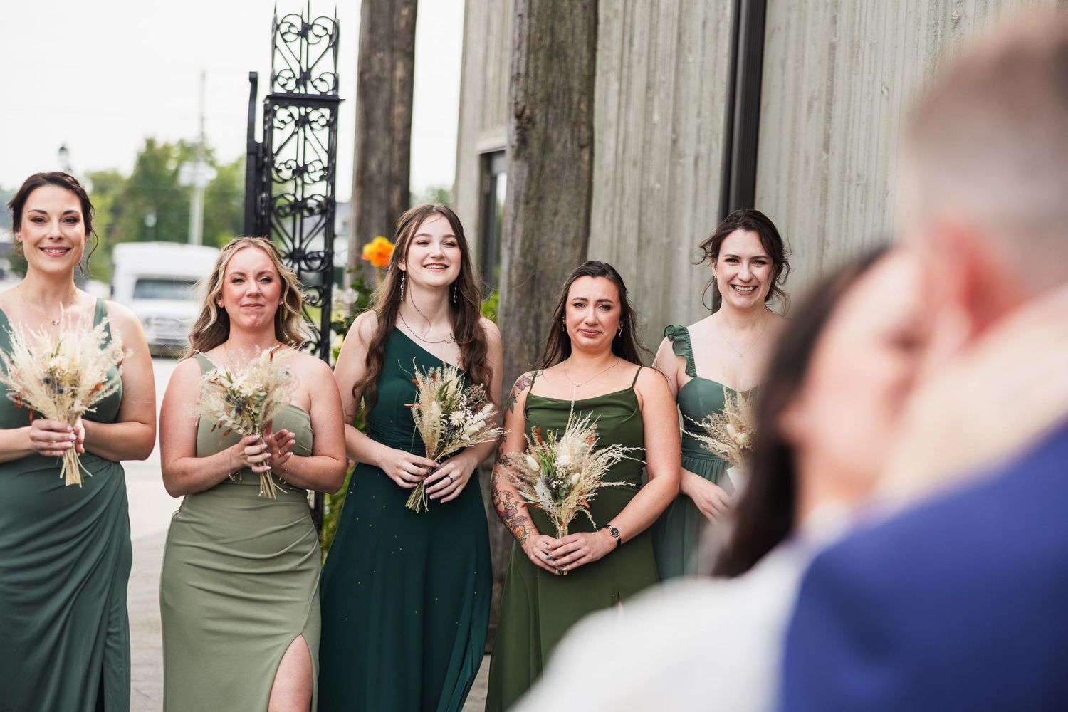 Wedding party of bridesmaids in varied green dresses smiling during outdoor ceremony.