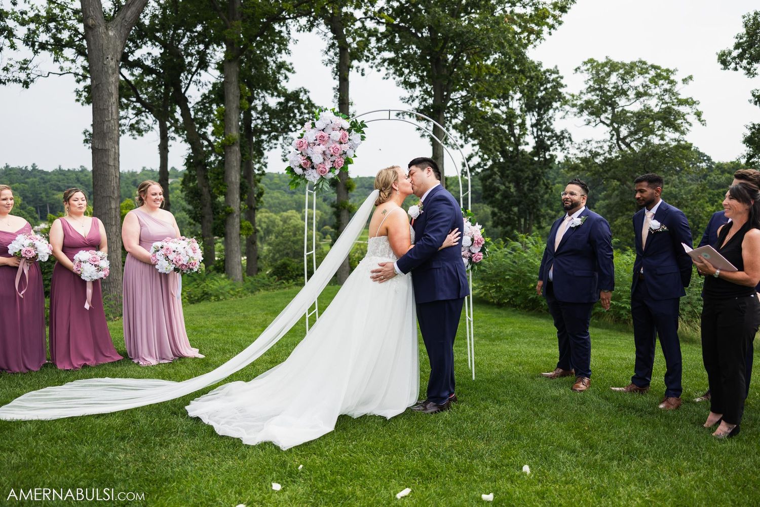 Wedding ceremony outdoors with bridesmaids in mauve dresses and groomsmen in navy suits under tall trees.