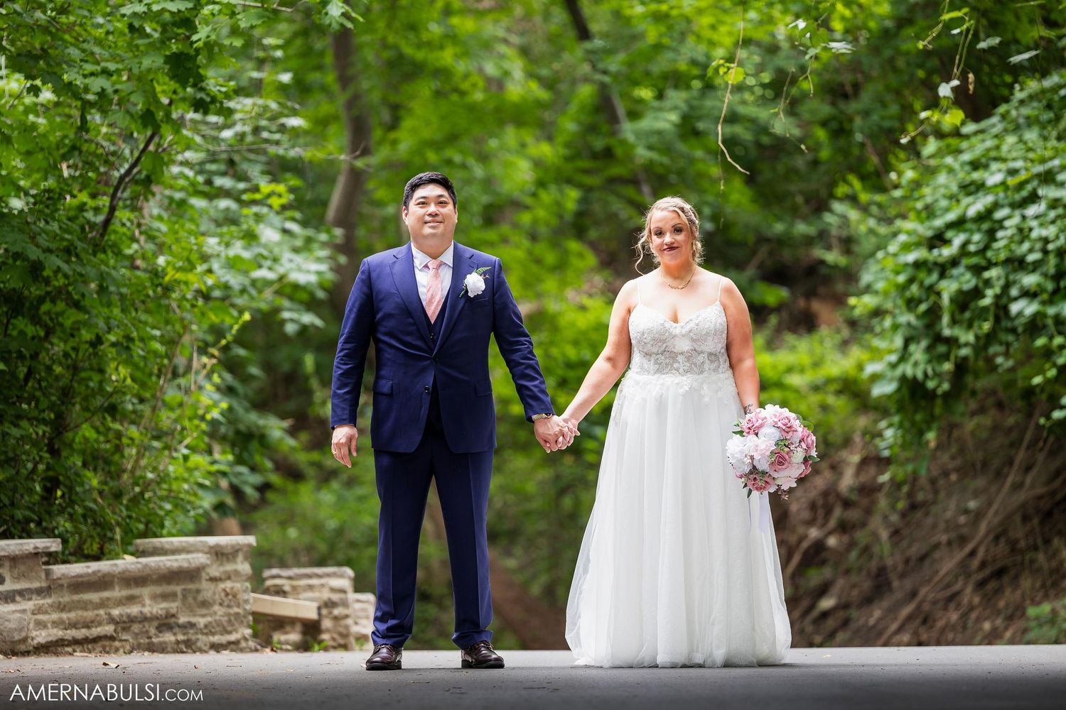 A wedding couple standing on a paved path surrounded by lush greenery.