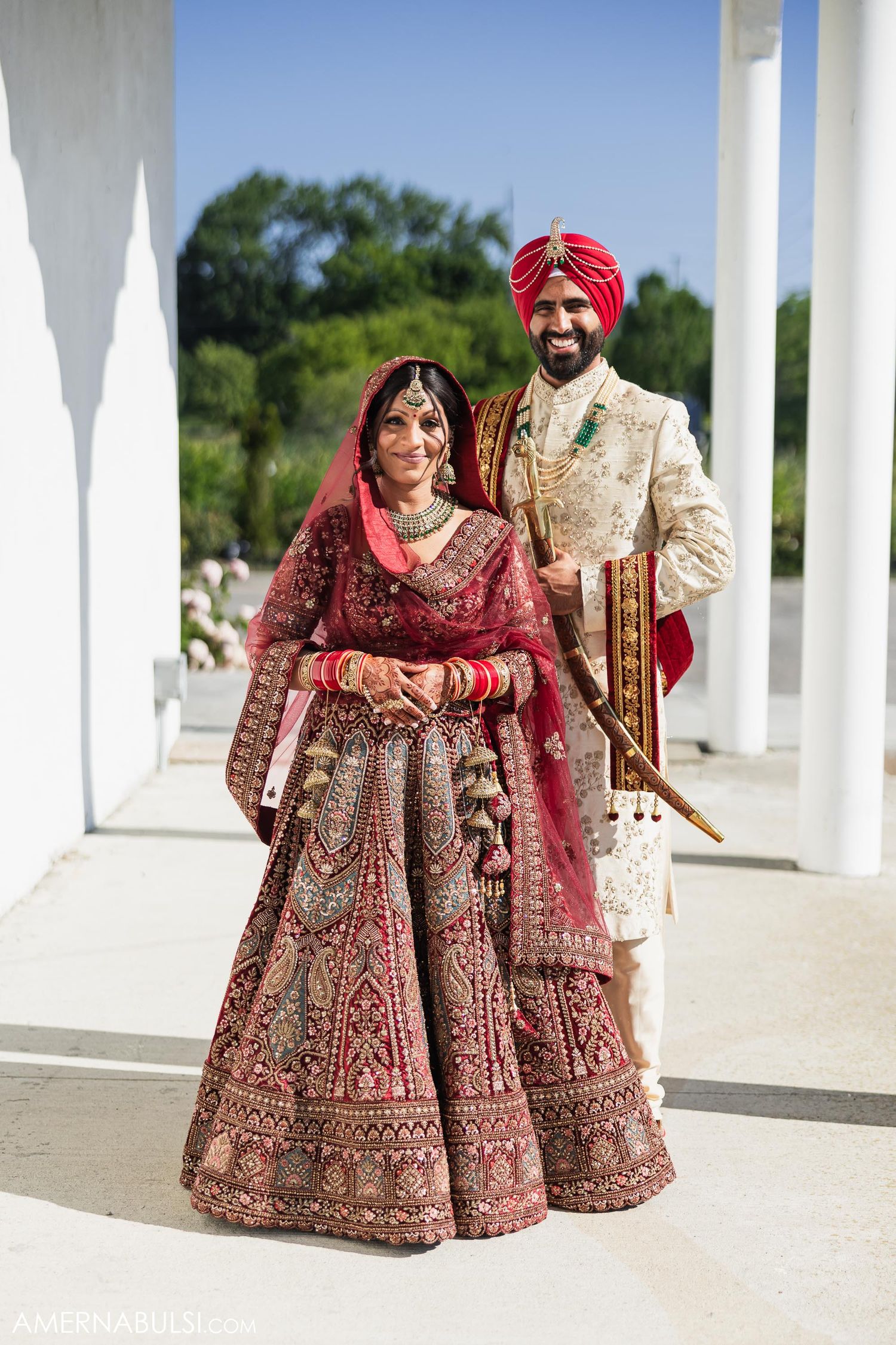 Traditional Indian wedding couple in ornate attire standing between white pillars.