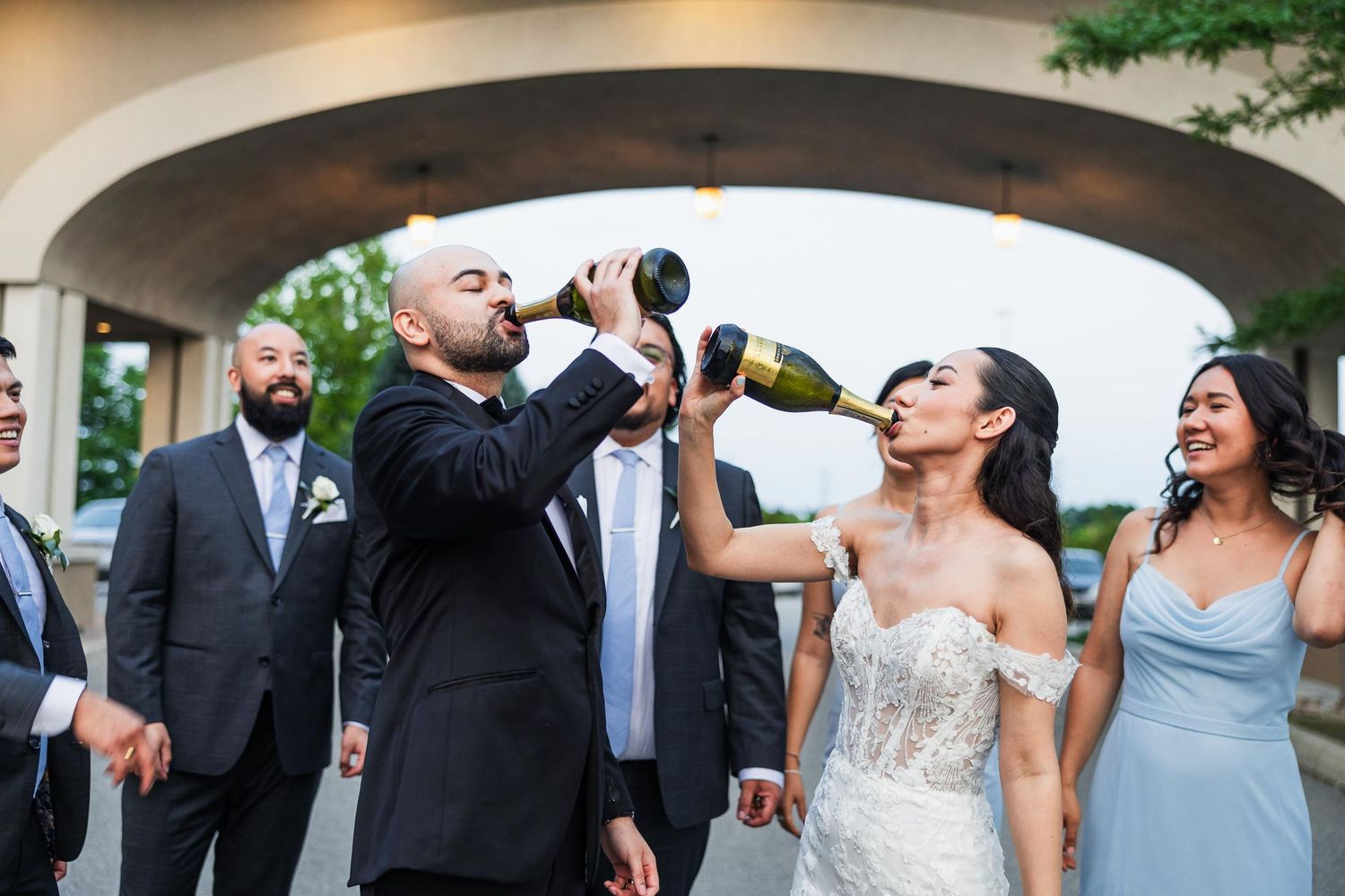 Wedding party members drink champagne under an outdoor archway during celebrations.