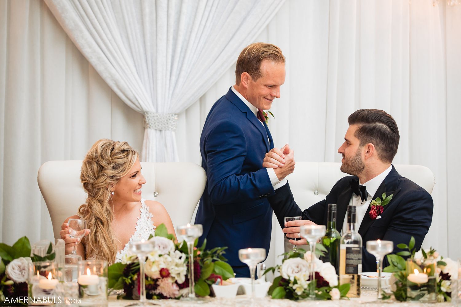 Wedding reception guests share joyful conversations and laughter at elegant white-draped tables with floral centerpieces.