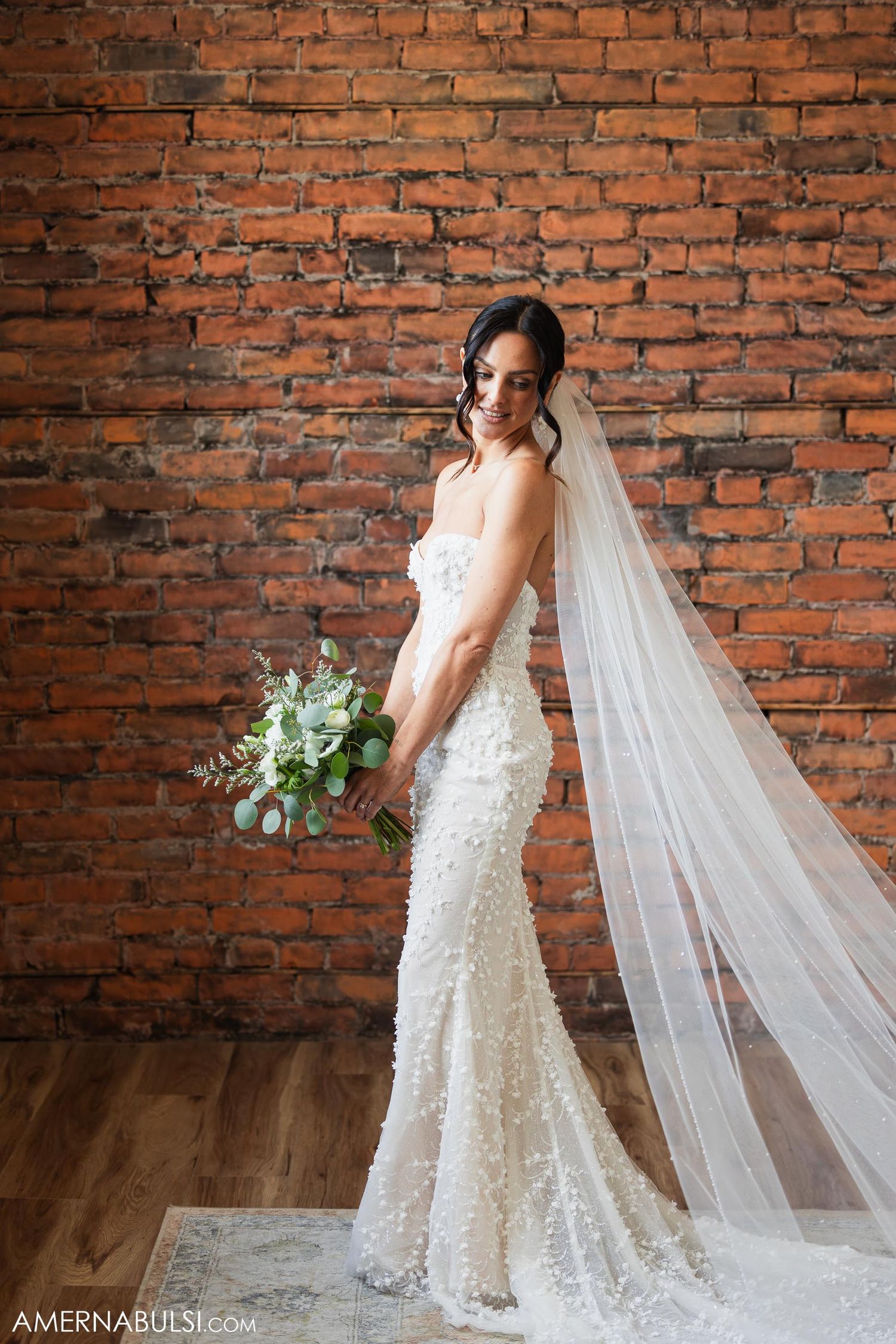 Bride in beaded form-fitting gown poses with bouquet against rustic brick backdrop.